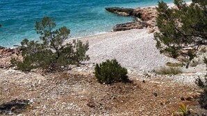 A coastal landscape with clear blue sea, rocky shoreline, and sparse vegetation.