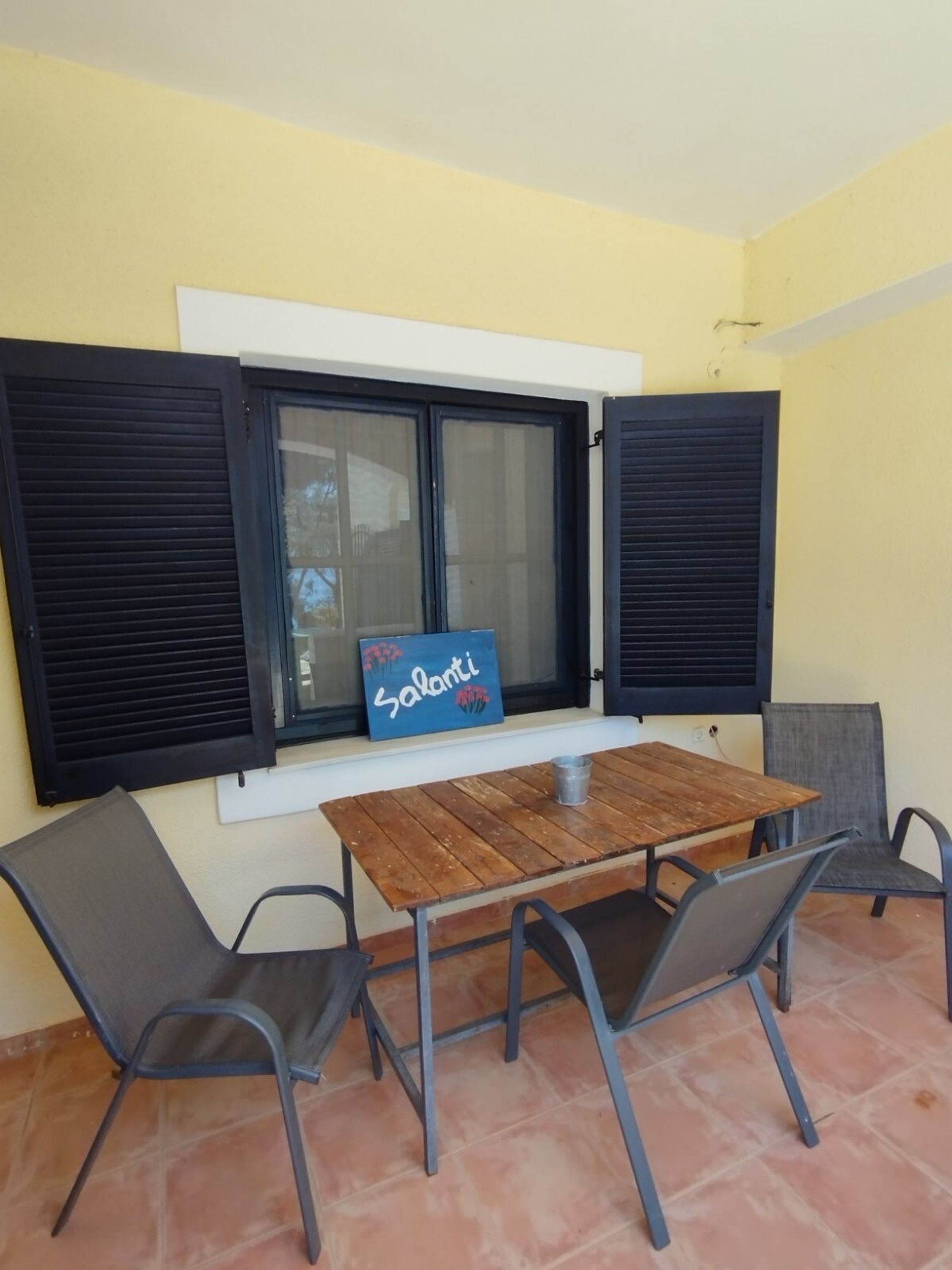 A small outdoor seating area with a wooden table and four chairs, a window with black shutters, and a sign reading "Salanti" on the window sill.