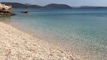 A pebble beach with clear water and distant mountains under a blue sky.
