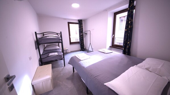 Un dormitorio con una cama nido, una cama individual, una mesita de noche, una ventana con cortinas y una lámpara de pie.