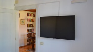 Een kamer met een wandgemonteerde flatscreen televisie, een boekenkast met boeken, een ingelijst schilderij aan de muur en een houten stoel.