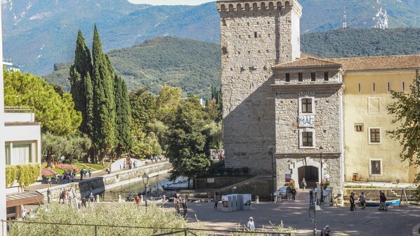 A historic stone tower with a flag, surrounded by lush greenery and a paved area with people and bicycles.
