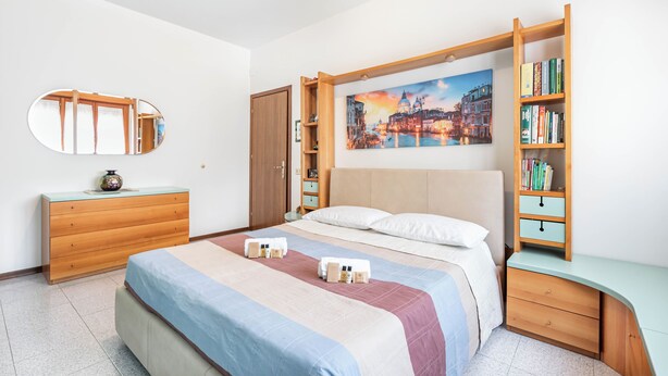 Ein Schlafzimmer mit einem Bett, einer hölzernen Kommode, einem Bücherregal, einem Spiegel und einem Stadtbild an der Wand.