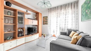 Ein modernes Wohnzimmer mit einem grauen Sofa, Holzregalen mit Glastüren, einem Flachbildfernseher und einer dekorativen Deckenleuchte.