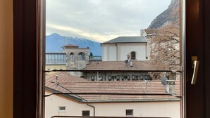 A view from a window overlooking a building with a tiled roof, a bell tower, and a mountain range in the background. - Two-Bedroom Apartment (Riva del Garda)