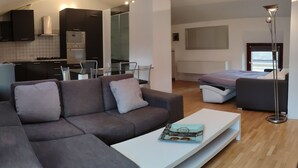 A modern living room with a grey sofa, a white coffee table, and a bed with a grey mattress. There is a kitchen area with dark cabinets and a white stove. - Two-Bedroom Apartment (Riva del Garda)