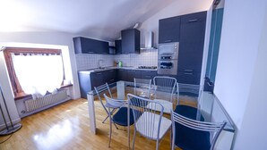 A modern kitchen with dark cabinets, a stainless steel oven, and a glass dining table with metal chairs. - Two-Bedroom Apartment (Riva del Garda)