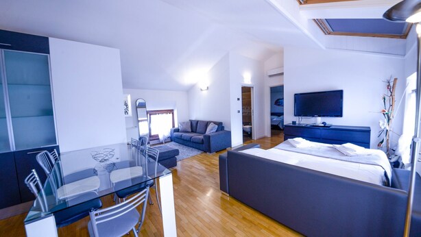 A modern hotel room with a glass dining table, a sofa, a bed, and a TV. - Two-Bedroom Apartment (Riva del Garda)