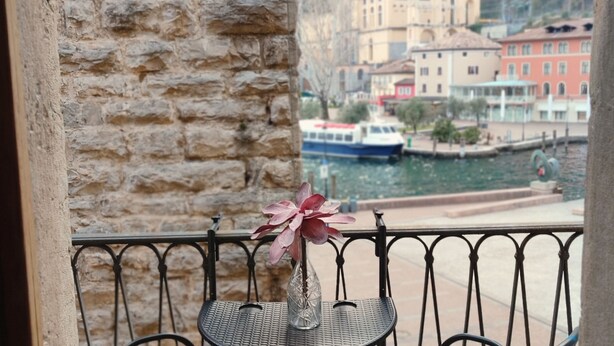 A balcony with a stone wall, a metal railing, and a table with a vase of flowers. In the background, there is a body of water with boats and buildings. - One Bedroom Apartment (Riva del Garda)