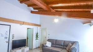 Un salon doté d’un téléviseur à écran plat, d’un canapé gris, de poutres en bois au plafond et d’une porte donnant sur une autre pièce.