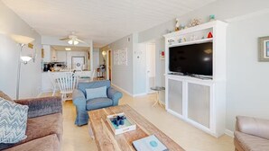 A living room with a TV, a coffee table, and a sofa. There is a kitchen in the background.