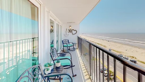 A balcony with a view of the beach, featuring a table with chairs and potted plants.