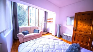 Une chambre à coucher avec un grand lit, un canapé, une armoire et une fenêtre donnant sur des immeubles et des arbres.