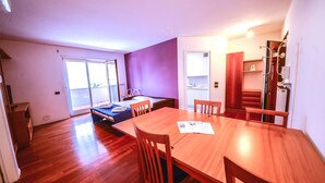 Une chambre d’hôtel moderne avec une table à manger en bois, un lit doté d’un matelas bleu et une porte-fenêtre coulissante donnant sur un balcon.