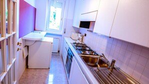 Une cuisine compacte avec des meubles blancs, une cuisinière à gaz, un évier et une fenêtre donnant sur un espace verdoyant.