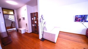 Una habitación moderna con piso de madera, una cama blanca, una mesita pequeña blanca, un armario de madera, una puerta de cristal y un televisor montado en la pared.