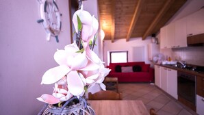 A living room with a red sofa, a kitchen area with cabinets and a countertop, and a vase with white flowers on a table. - Two-Bedroom Apartment (Arco)