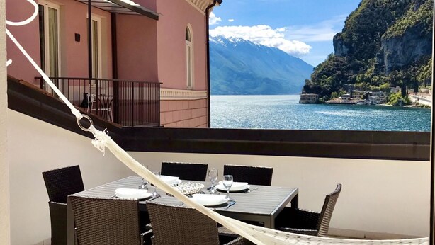 Un balcón con muebles de mimbre y una hamaca, con vista a un lago y montañas, con edificios al fondo.