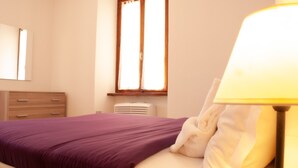 Een slaapkamer met een bed, nachtkastje, lamp, raam en een airconditioner.