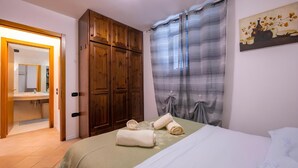 A bedroom with a bed, wooden wardrobe, bathroom visible through an open door, and a painting on the wall.