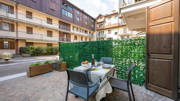 A courtyard with a dining table set for two, surrounded by a green hedge, with multi-story buildings in the background.