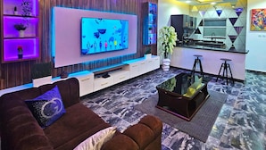 Una sala de estar moderna con un televisor grande, una araña y una zona de cocina.