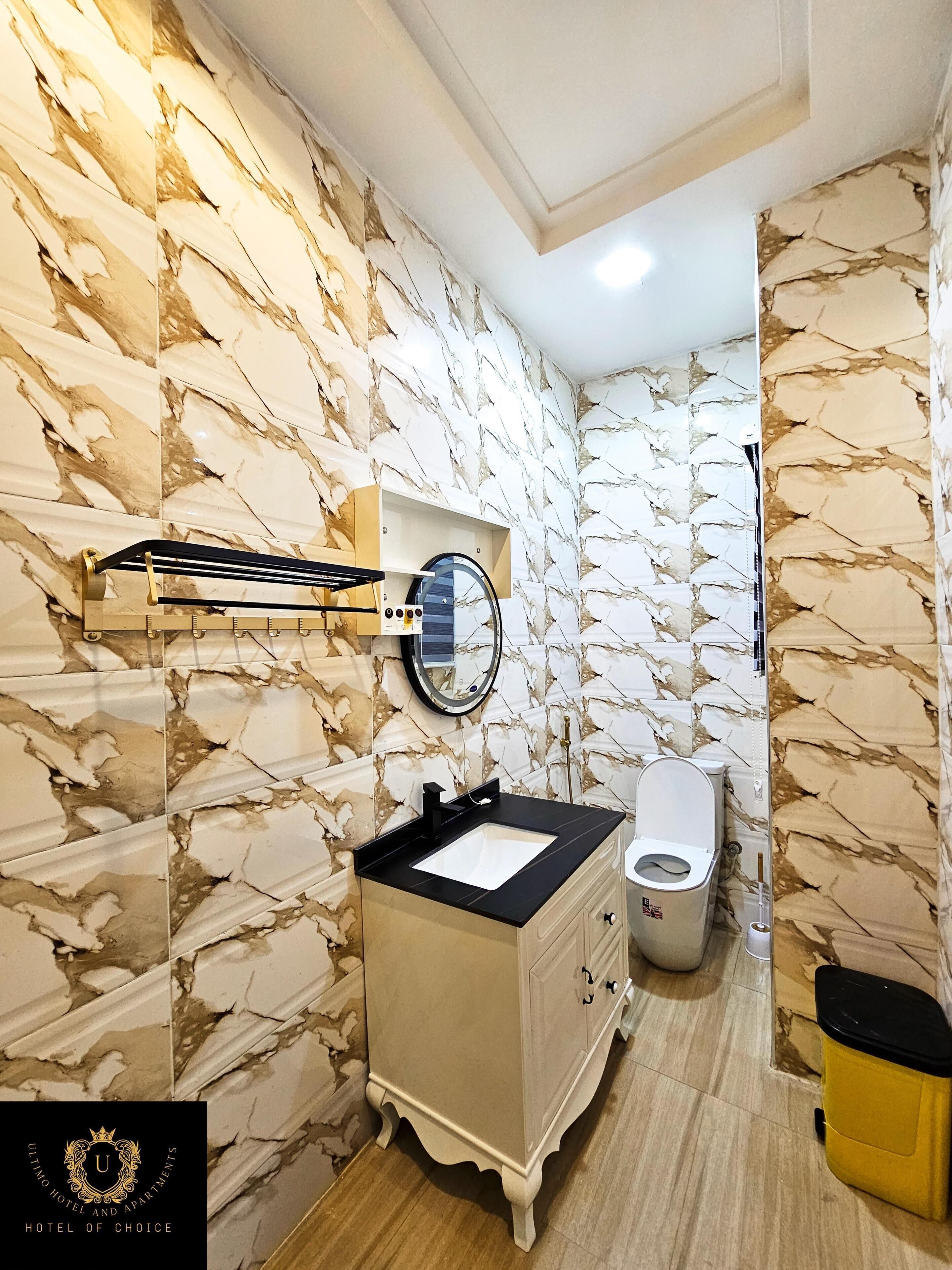 Un baño con paredes de diseño de mármol, un tocador con lavabo, un espejo, un inodoro y un cubo de basura.