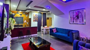 A modern living room with a blue sofa, a coffee table, and a TV. There is a dining area with a table and chairs. - 2 Bed Apartment 1 - Stylish Benin City Retreat (Benin City)