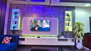 A modern living room with a large flat-screen TV mounted on the wall, a blue sofa with patterned cushions, a wooden coffee table, and a red carpet. - 2 Bed Apartment 1 - Stylish Benin City Retreat (Benin City)