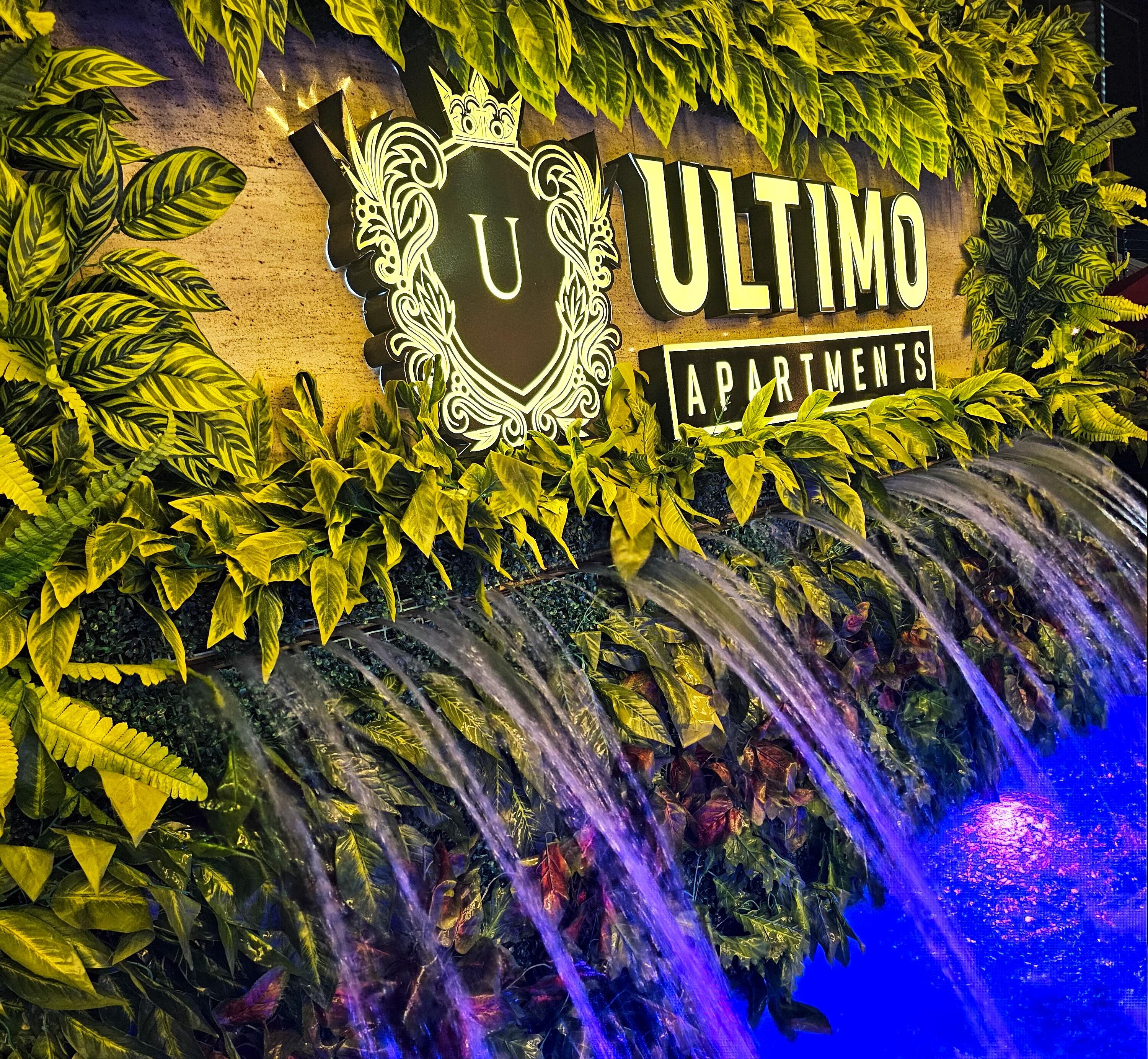 A sign for Ultimo Apartments with a logo and text, surrounded by lush greenery and a water feature.