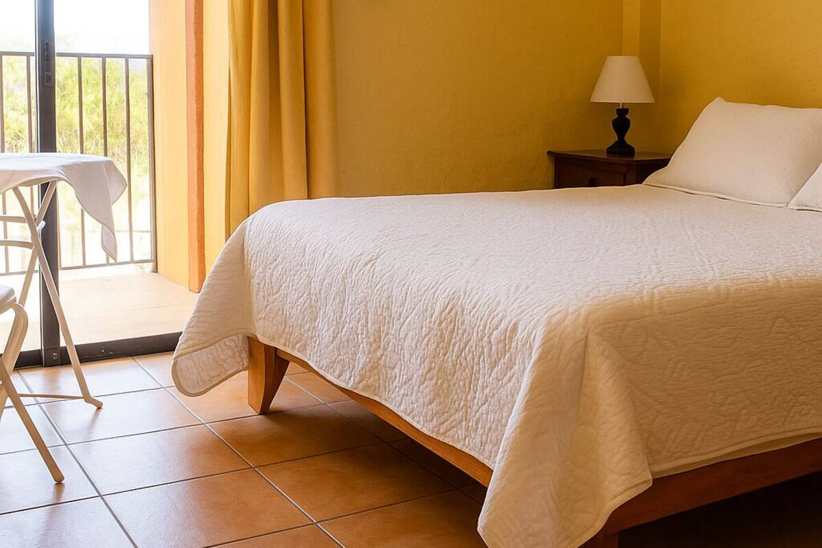 Ein Schlafzimmer mit einem Bett, einem Nachttisch, einer Lampe, einem Sessel und Blick ins Freie durch eine Schiebetür.