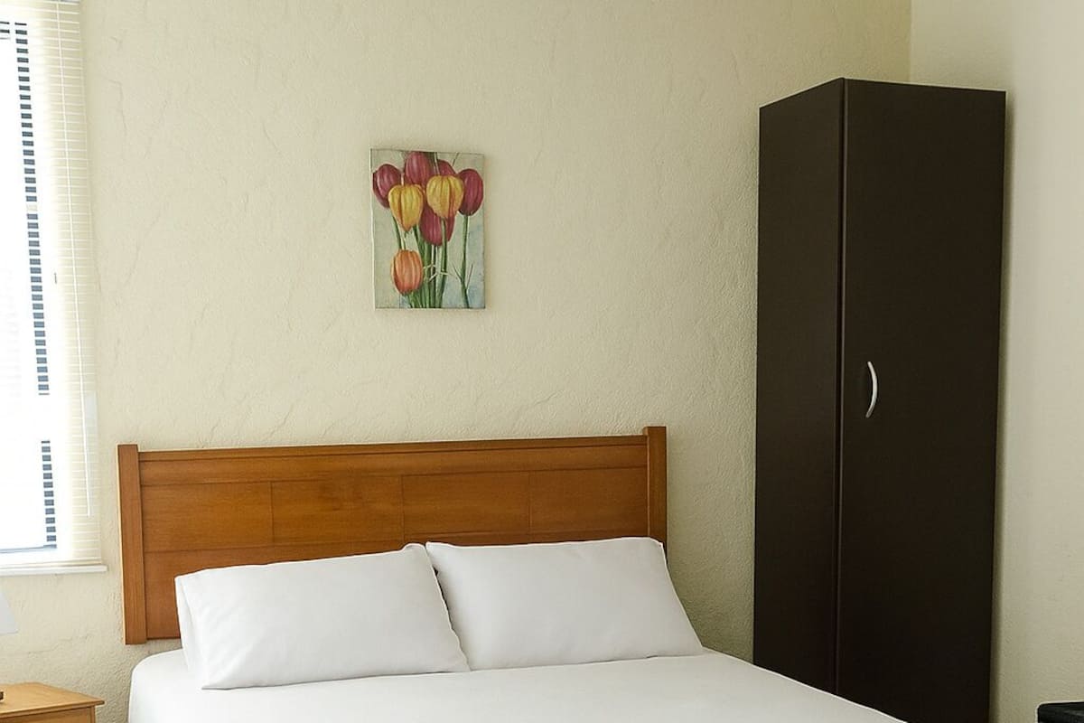 Ein Schlafzimmer mit einem hölzernen Kopfteil, weißer Bettwäsche, einem Nachttisch, einem Kleiderschrank und einem Gemälde von Tulpen an der Wand.