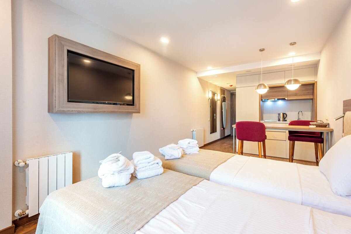 Ein Hotelzimmer mit zwei Betten, einem an der Wand befestigten Fernseher, einem kleinen Schreibtisch mit Stuhl und einem durch eine offene Tür einsehbaren Badezimmer.