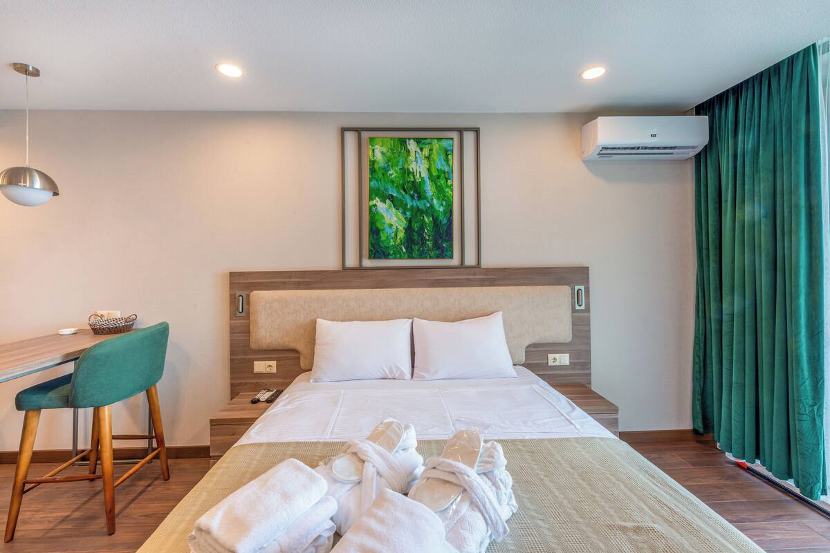 Ein Hotelzimmer mit einem Bett, einem Schreibtisch mit Stuhl, einem gerahmten Gemälde, einer Klimaanlage und einem Fenster mit Vorhängen.