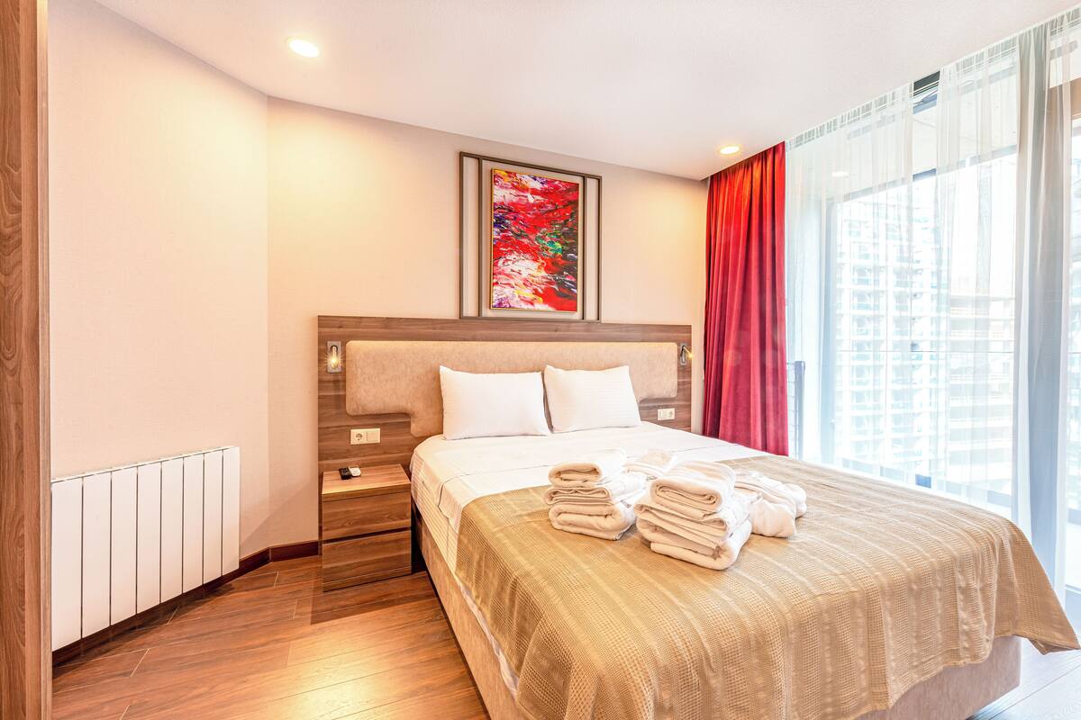 Ein Hotelzimmer mit einem großen Bett, einem Nachttisch, einem Fenster mit Vorhängen und einem gerahmten Kunstwerk an der Wand.