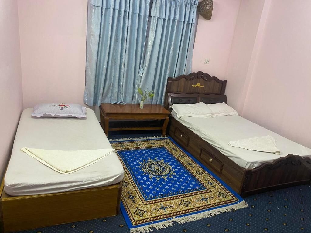 A small, simple room with two single beds, a wooden nightstand, a blue patterned rug, and a window with light blue curtains.