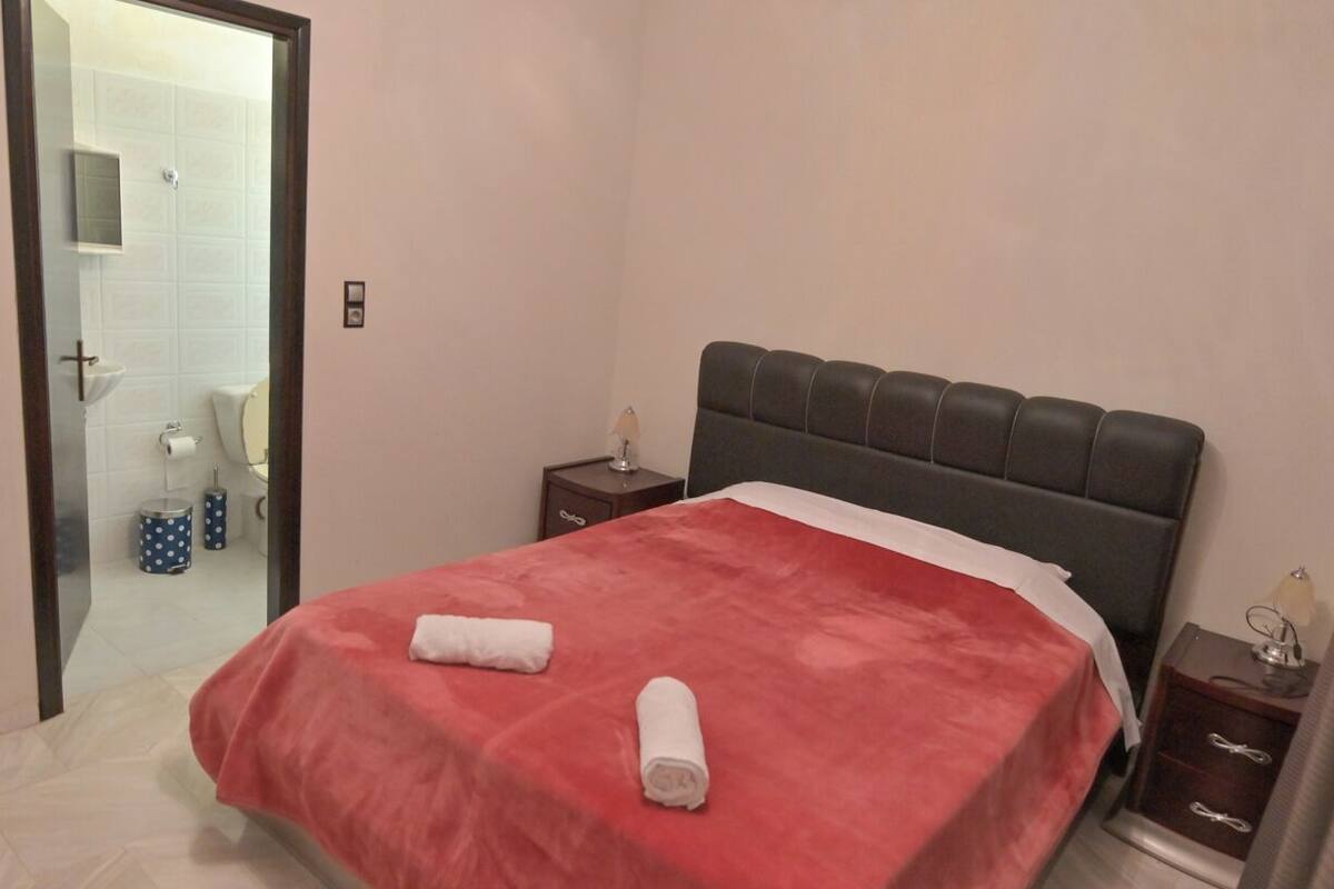 Una habitación de hotel con cama, mesita de noche, lámpara y baño visible a través de una puerta abierta.