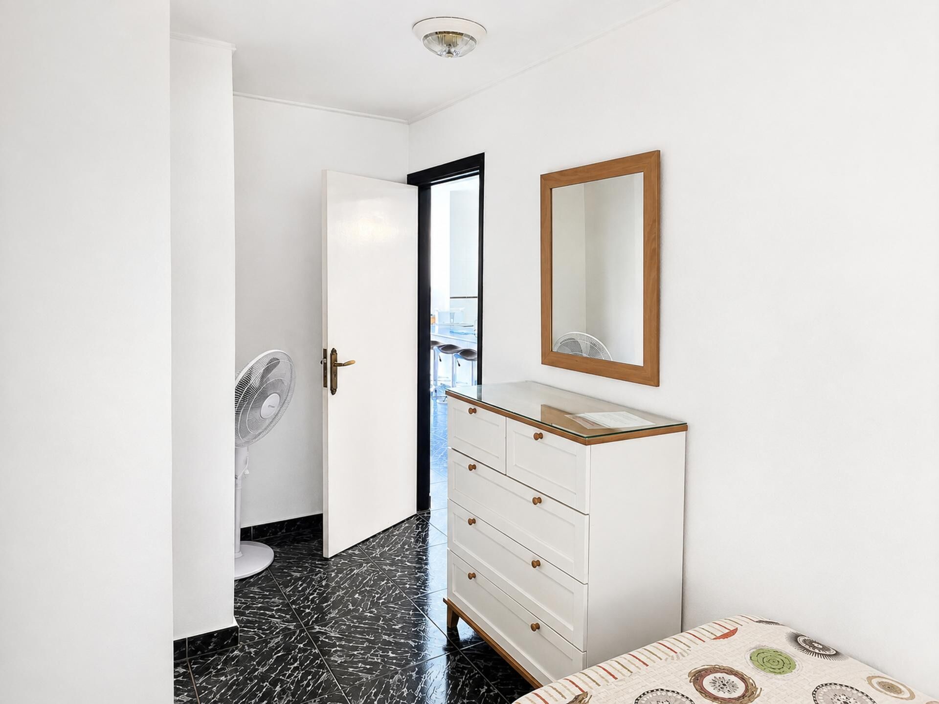 Une chambre d’hôtel avec une armoire blanche, un miroir, un ventilateur et une couvre-lit à motifs.