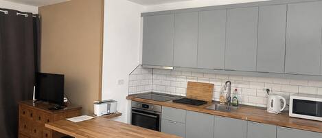 A compact kitchen with grey cabinets, a wooden countertop, a built-in oven, a microwave, and a small sink. There are two black bar stools and a television on a wooden cabinet.