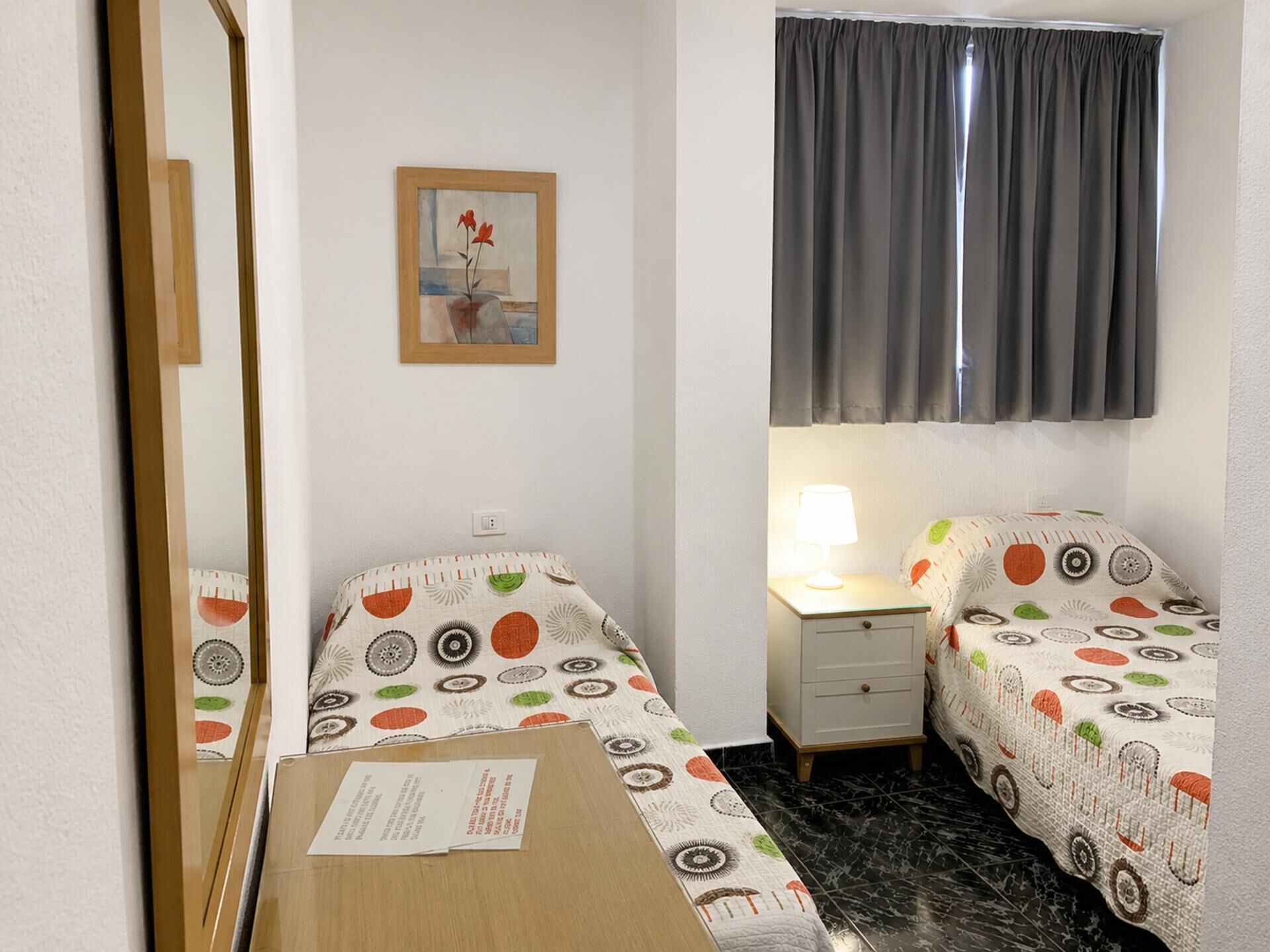 A small room with two single beds, a desk, a lamp, a mirror, and a framed picture on the wall.