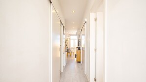 A narrow hallway with white walls and a light wooden floor. There are doors on both sides and a small table with chairs in the distance.
