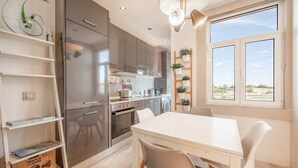 A modern kitchen with stainless steel appliances, a dining area with white furniture, and a view of the outdoors through large windows.