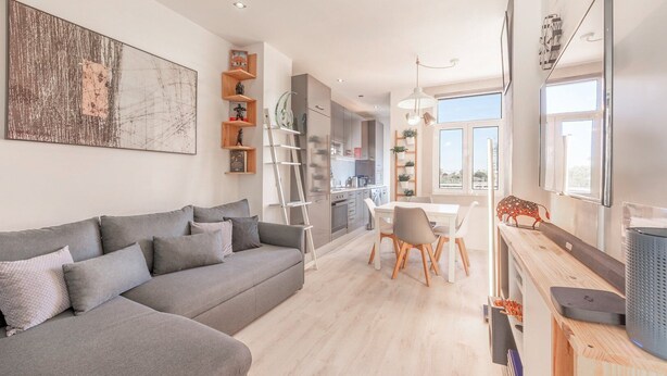 A modern living room with a grey sofa, a dining area with a white table and chairs, and a kitchen with wooden cabinets and a ladder.