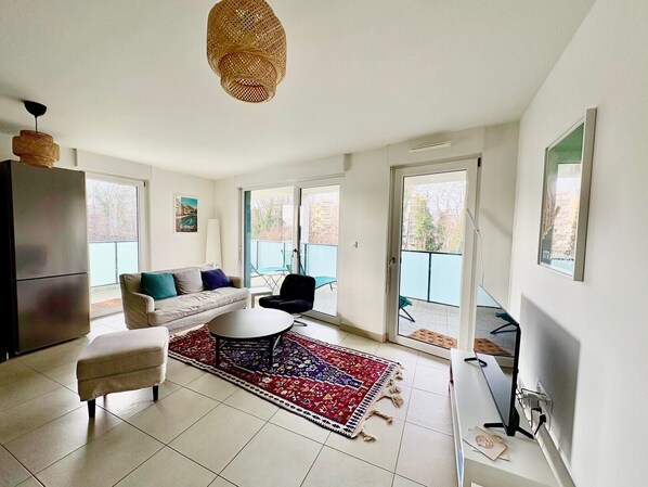A modern living room with a sofa, a coffee table, and a rug. There is a balcony with a blue railing and a chair.