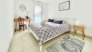Een slaapkamer met een bed, een bureau, een stoel, een nachtkastje, een lamp, een spiegel, een schilderij aan de muur en een raam met uitzicht.