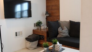 Un salon moderne avec un téléviseur à écran plat fixé au mur, un mur d’accent en bois, un canapé de couleur foncée agrémenté de coussins, une petite table d’appoint avec une lampe, une plante en pot et une télécommande posée sur une table blanche.