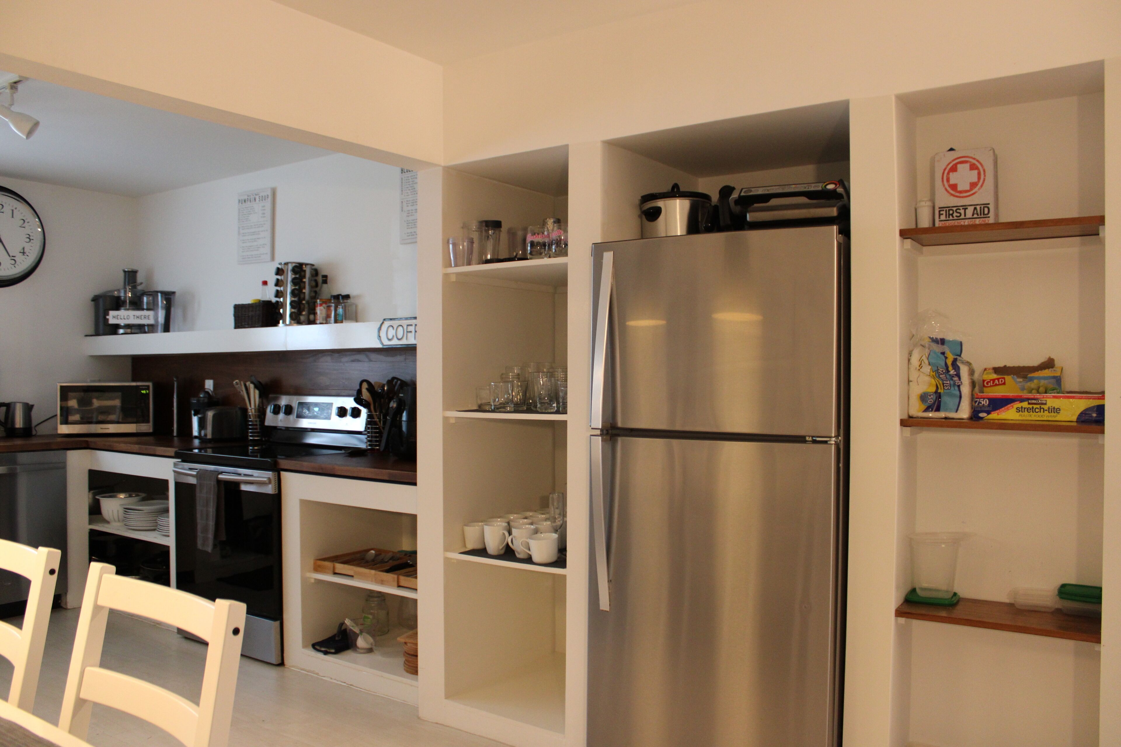 A compact kitchen with a stainless steel refrigerator, built-in oven, and microwave. There are white cabinets and shelves with various kitchen items.