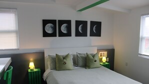 Une chambre à coucher avec un lit, des tables de chevet vertes, un bureau et une chaise. Il y a quatre tableaux représentant les différentes phases de la lune accrochés au mur.