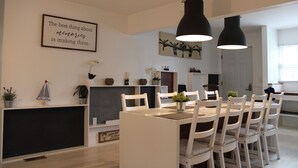 Un espace repas moderne avec une table et des chaises blanches, des suspensions noires et une citation encadrée au mur.