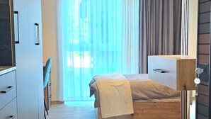 A modern hotel room with a wooden floor, a bed with a white cover, a desk with a chair, a large window with sheer curtains, and a bright yellow pendant light. - Hira Boarding Aparthotel (Dingolfing)